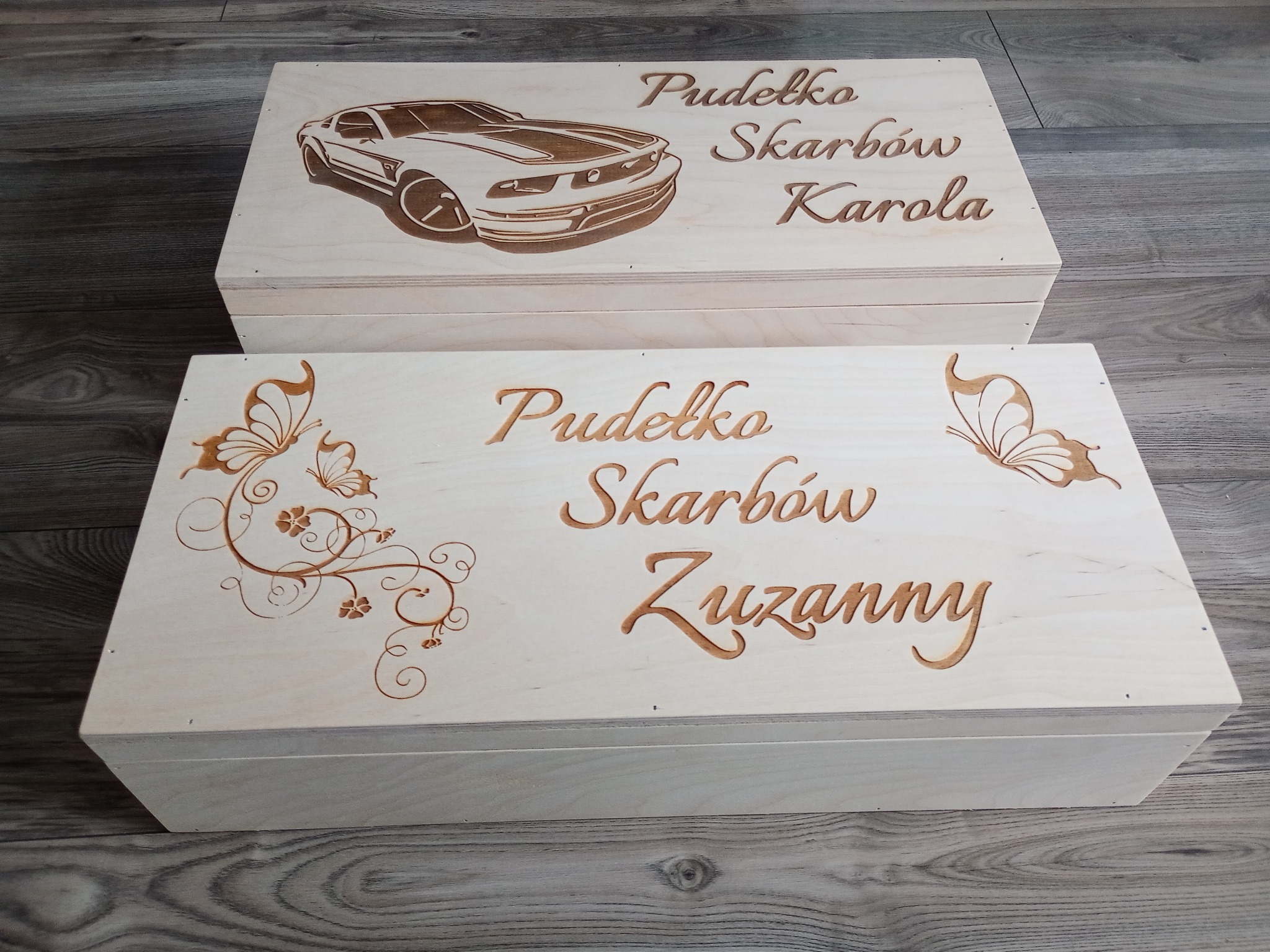 Dwa drewniane pudełka z wygrawerowanymi wzorami: samochód i motyle, oraz napisy 'Pudełko Skarbów Karola' i 'Pudełko Skarbów Zuzanny'.