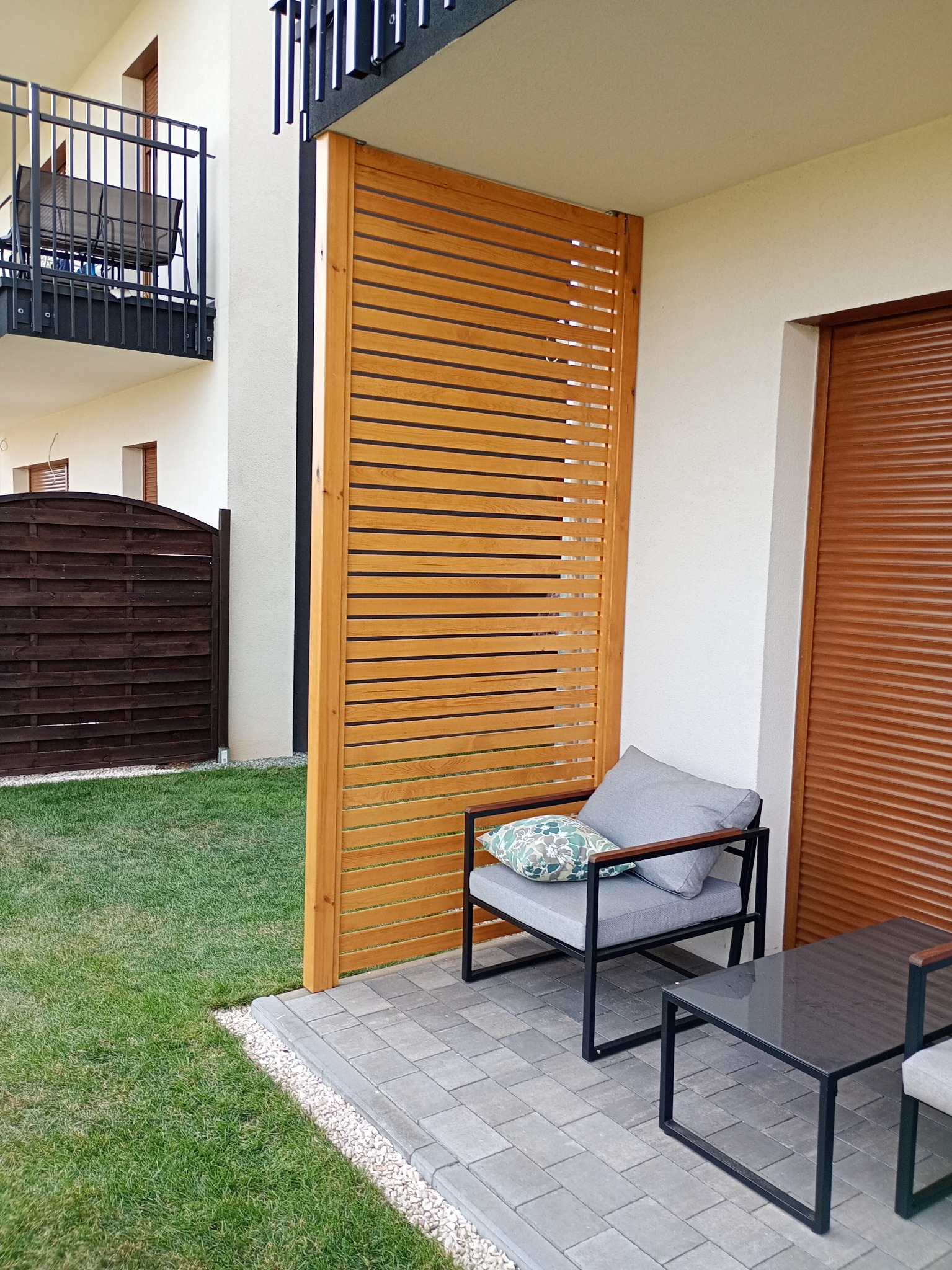 Nowoczesny taras z drewnianą przesłoną, szarą sofą i stolikiem kawowym na tle zielonego trawnika i drewnianego ogrodzenia. Ujęcie z niskiego kąta.