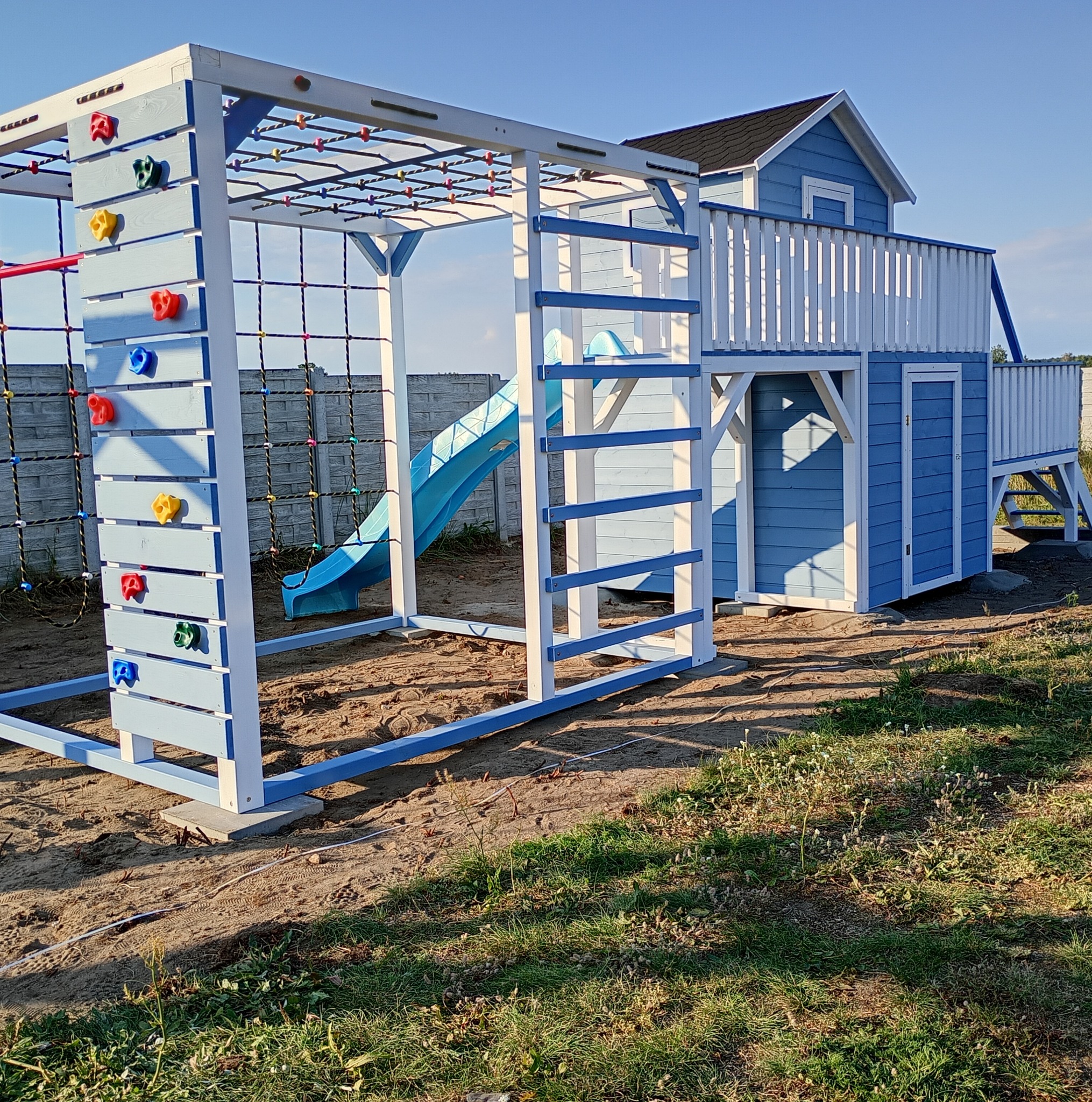 Niebiesko-biały plac zabaw z domkiem, zjeżdżalnią i ścianką wspinaczkową na piaszczystym podłożu, w tle szary betonowy płot i błękitne niebo.