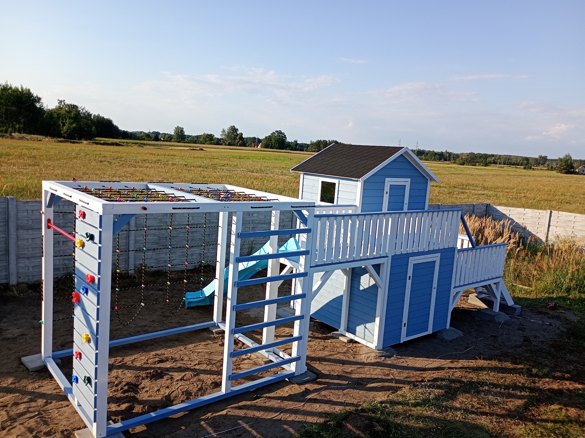 Niebiesko-biały plac zabaw z domkiem, drabinką i ścianką wspinaczkową na tle pola i błękitnego nieba. Widok z perspektywy dziecka, w słoneczny dzień.