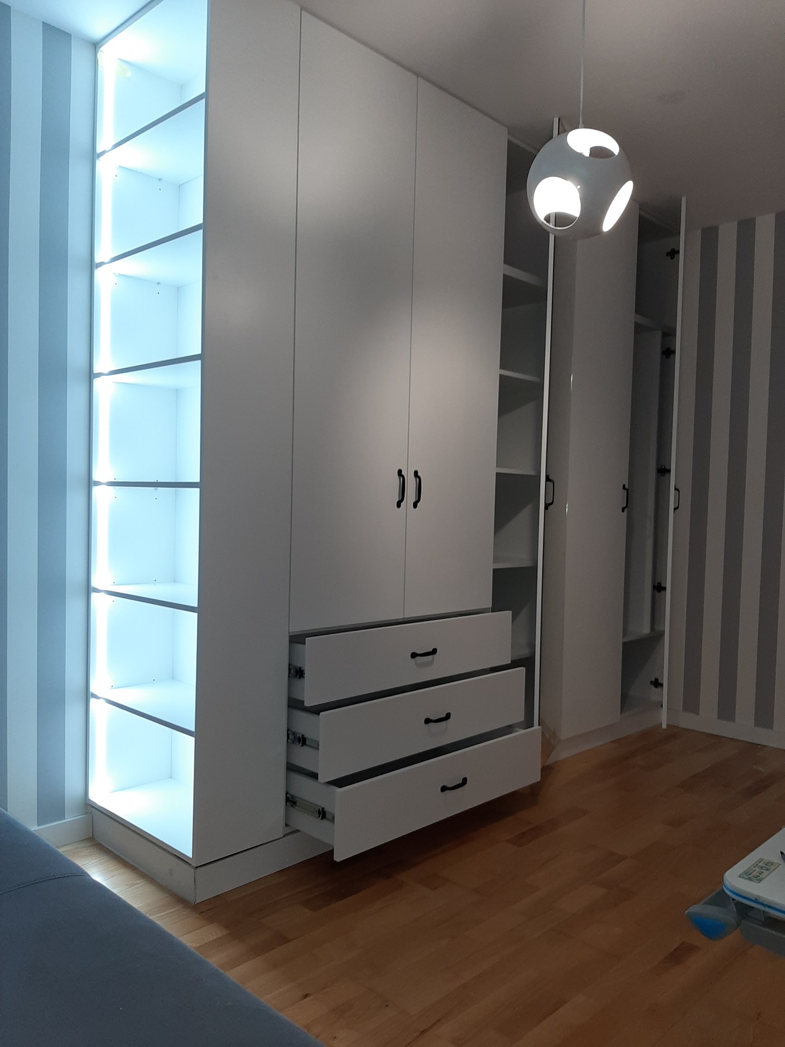 Biała szafa wnękowa z oświetleniem LED i wysuwanymi szufladami, widoczna na tle ściany w paski. Nowoczesny design, minimalistyczny styl, drewniana podłoga.