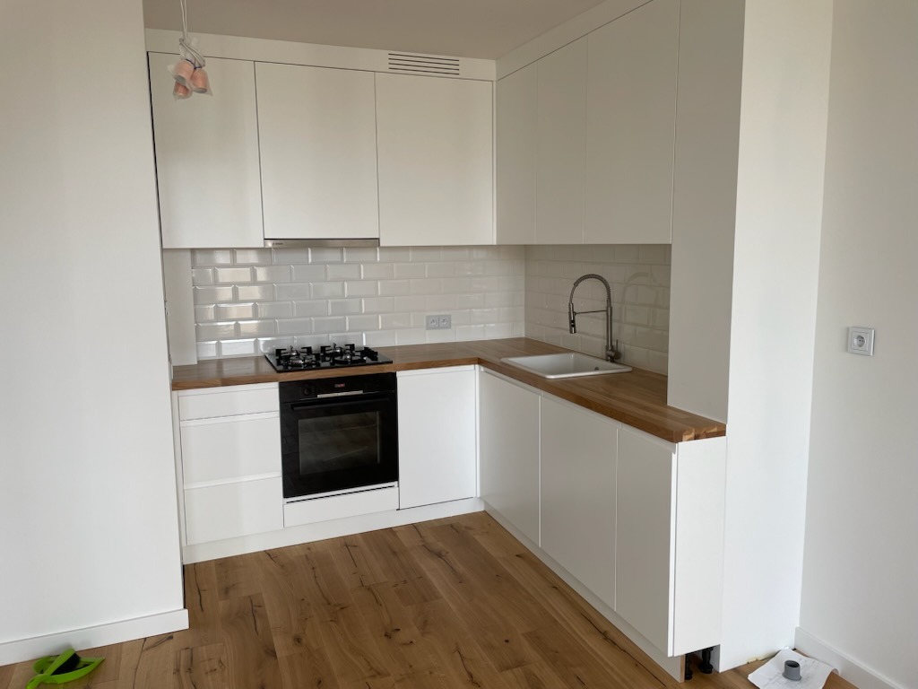 Biała, narożna kuchnia z drewnianym blatem i białymi płytkami na ścianie. Widoczny zlew, płyta grzewcza i piekarnik. Podłoga drewniana. Minimalistyczny design.