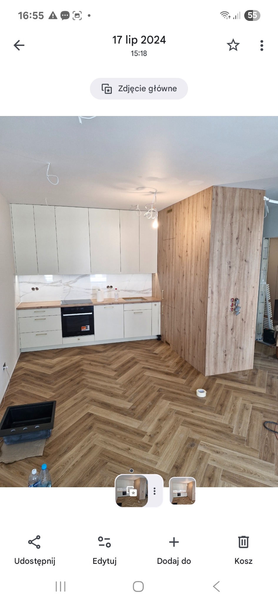 Jasna kuchnia z drewnianą podłogą w jodełkę i białymi szafkami. Widoczny fragment ściany z drewnianą okładziną. Uwagę zwraca nowoczesny design i dbałość o detale.