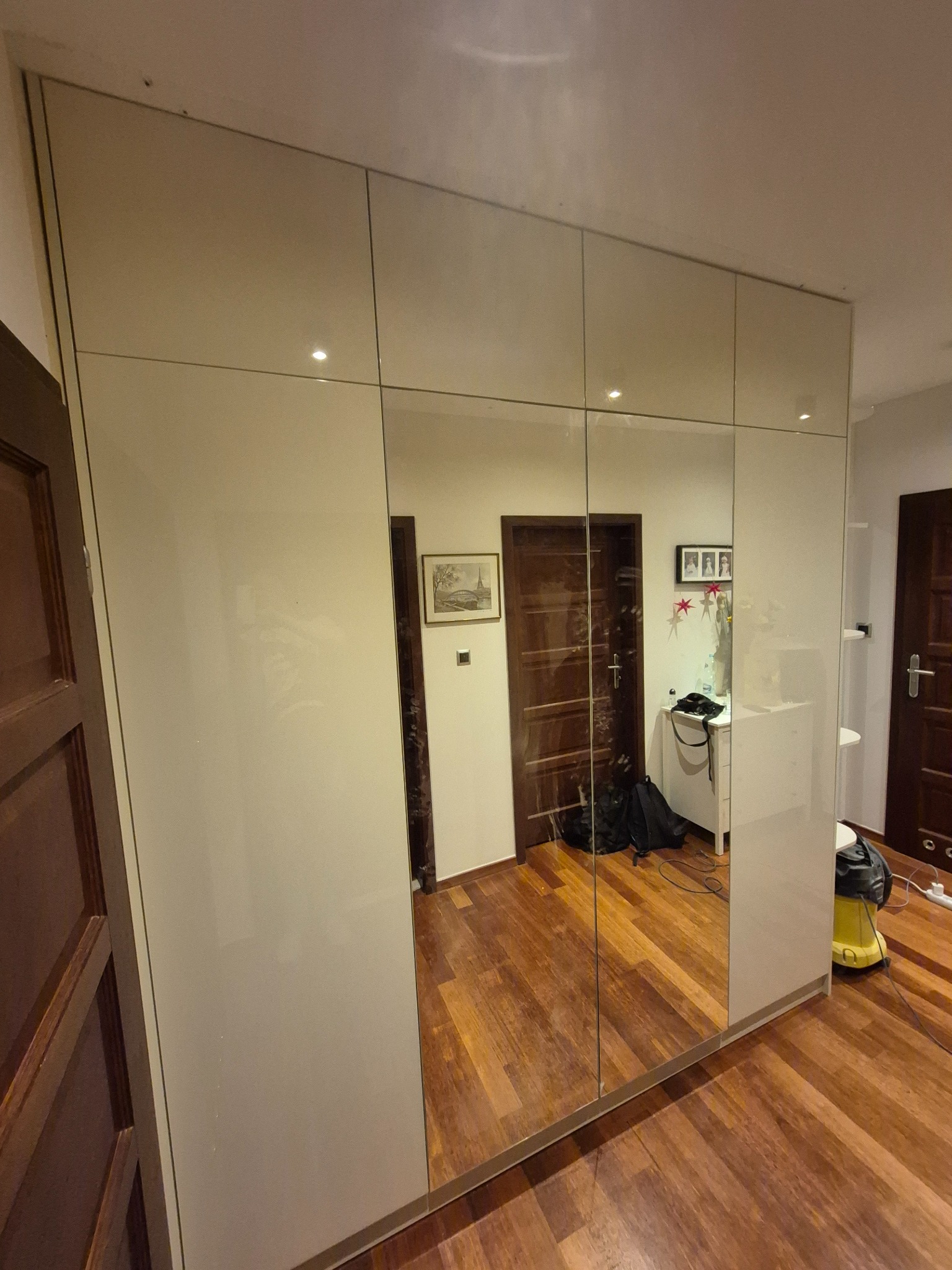 Biała szafa wnękowa z lustrzanymi drzwiami, odbijająca wnętrze pokoju z drewnianą podłogą i elementami wyposażenia. Widoczne oświetlenie punktowe.