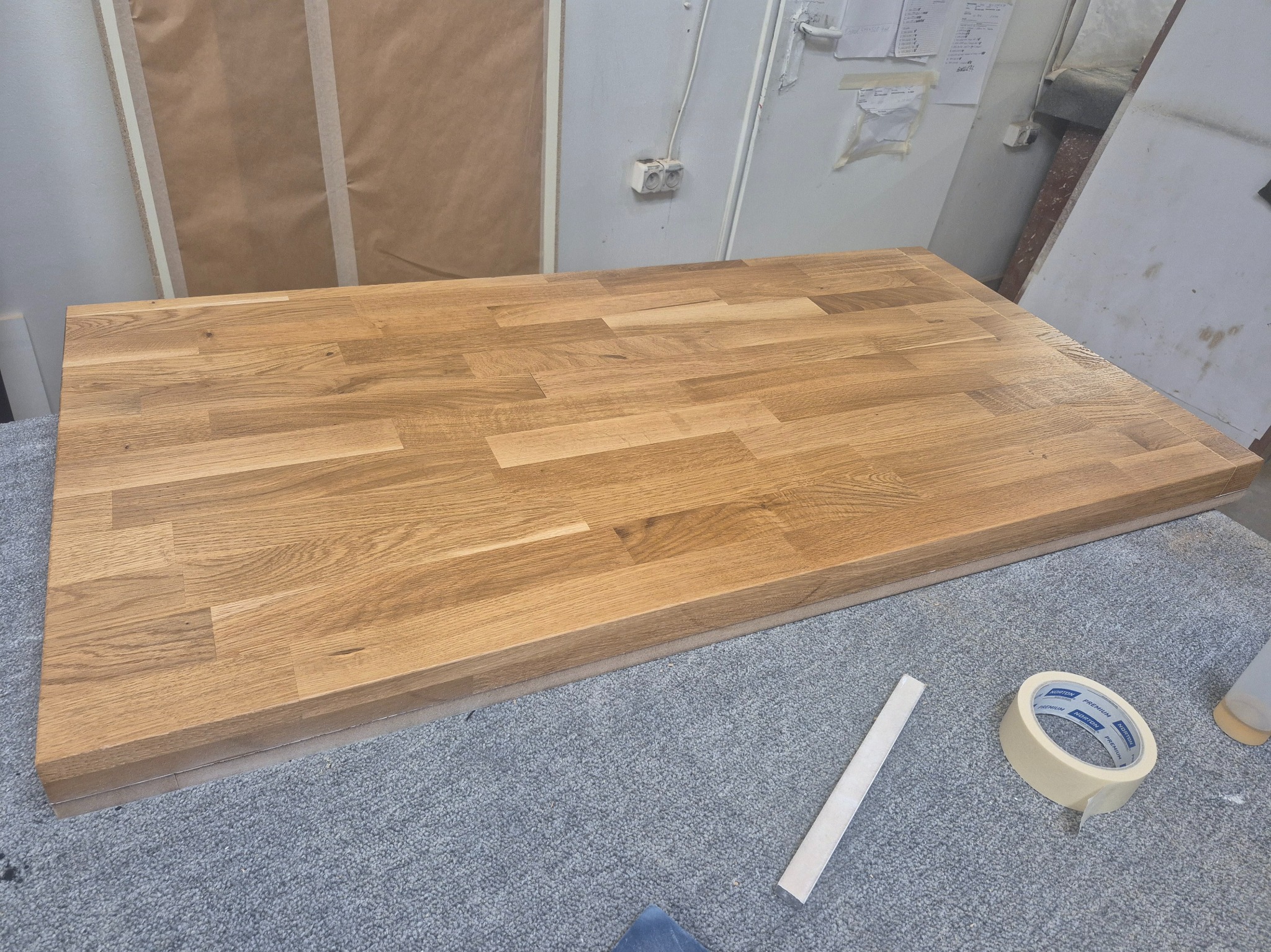 Blat dębowy po olejowaniu, leżący na szarym dywanie, obok taśma malarska i listwa. Widoczne gniazdka elektryczne na ścianie. Jasne, naturalne światło.