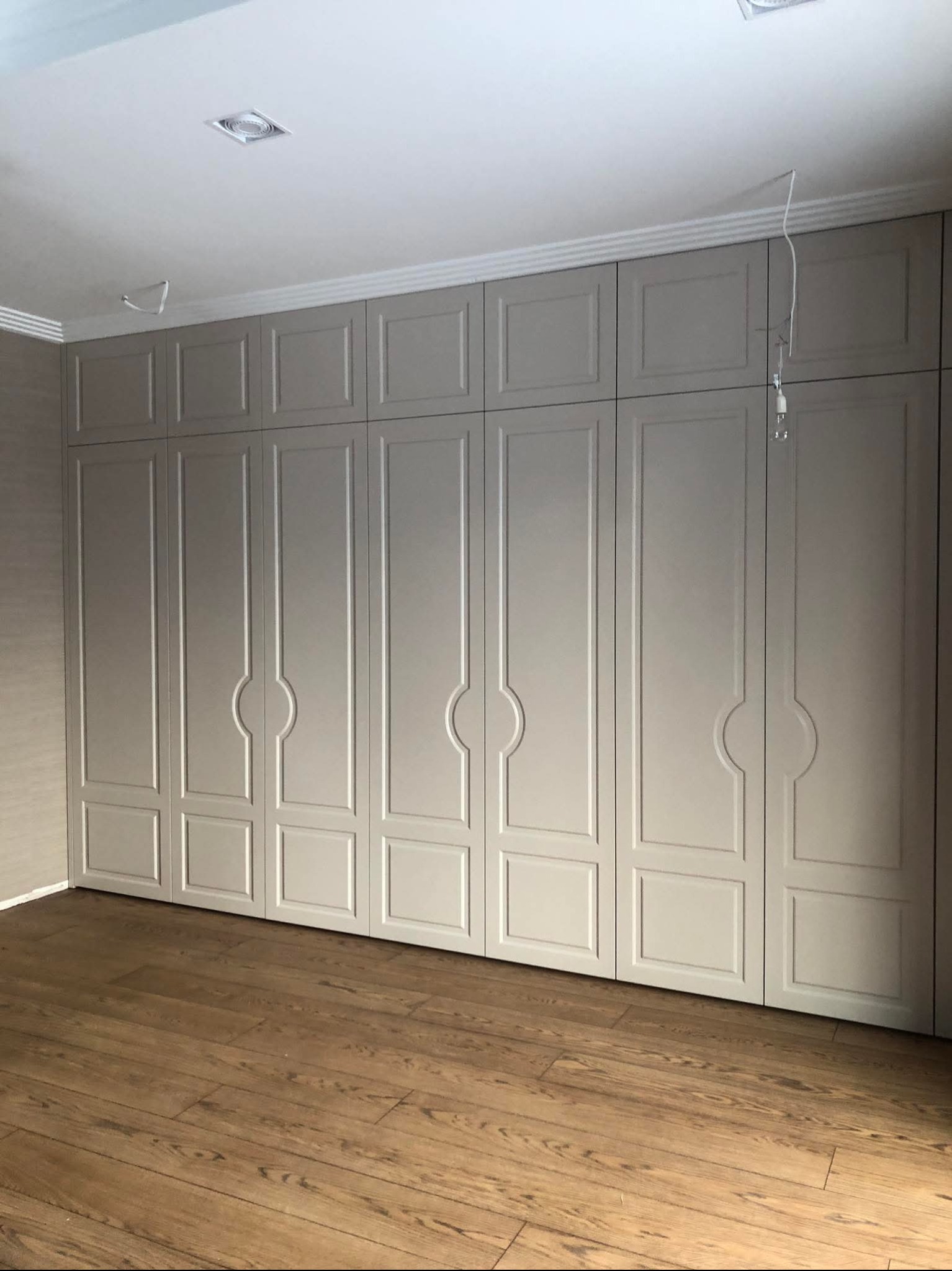 Jasnoszara szafa wnękowa z frezowanymi drzwiami i ozdobną listwą pod sufitem, na drewnianej podłodze. Widoczne niedokończone podłączenie oświetlenia.