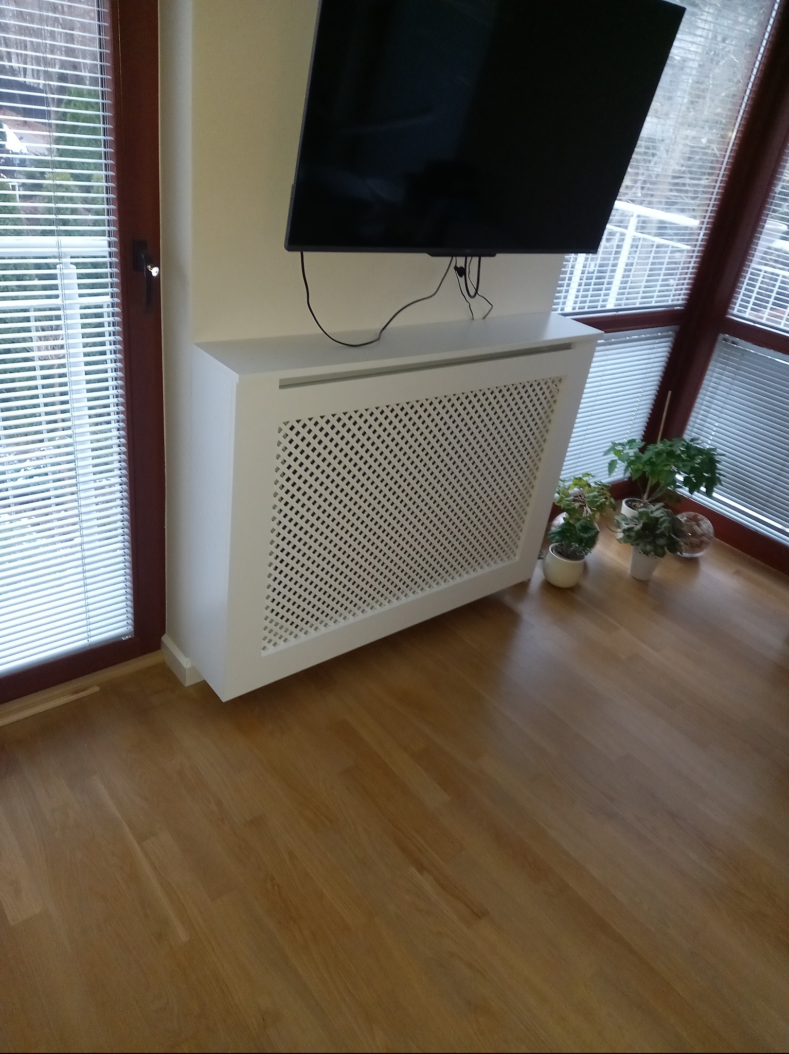 Biała obudowa grzejnika z ażurowym frontem, dopasowana do ściany i pod telewizorem. Obok rośliny doniczkowe i okno z żaluzjami. Parkiet na podłodze.