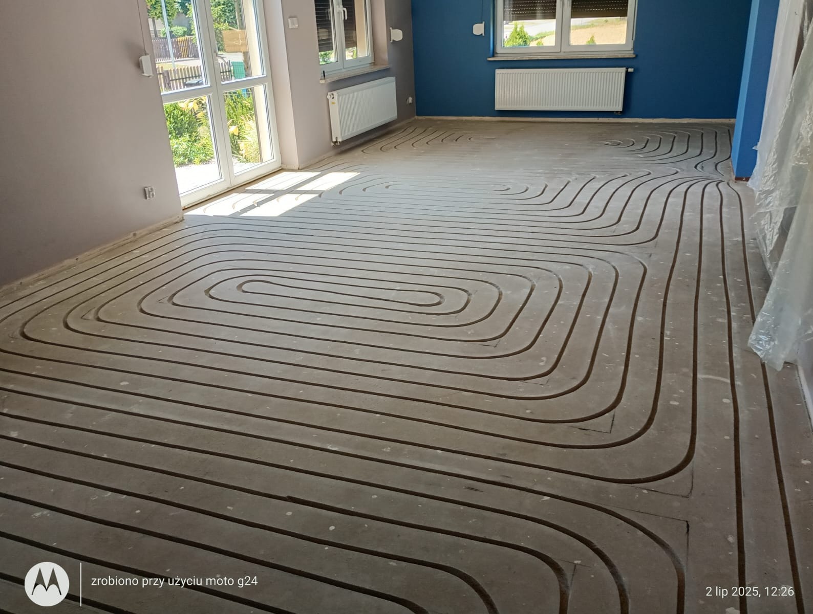 Ułożone rury ogrzewania podłogowego w nowym budynku, widok z góry. Równoległe, zakrzywione linie na szarej powierzchni. Świeże światło wpada przez okno.