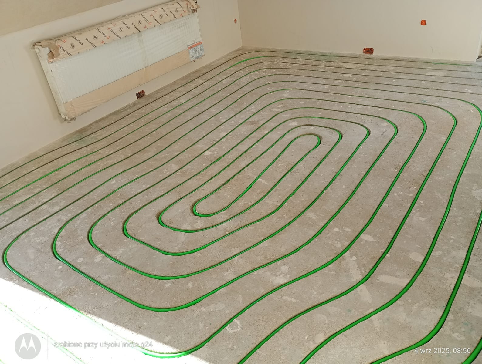 Zielone rurki ogrzewania podłogowego ułożone spiralnie na szarym betonie, grzejnik w folii na ścianie. Widok z góry, surowy stan przed wylewką.