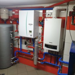 Winenergy - Zestaw urządzeń grzewczych: zbiornik, pompa ciepła Ecodan i piec Buderus, połączone rurami w kolorach niebieskim i czerwonym. Instalacja w pomieszczeniu z czerwono-białymi płytkami.