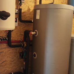 Winenergy - Instalacja CO z zbiornikiem buforowym, rurami w izolacji i zaworami, zamontowana na ścianie z boazerii. Widoczne elementy instalacji grzewczej.