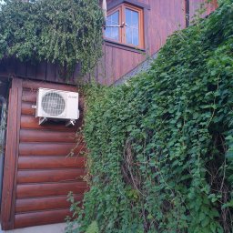 Winenergy - Klimatyzator Mitsubishi Electric zamontowany na drewnianej elewacji budynku, częściowo porośniętej zielenią. Widoczny fragment okna i dachu. Ujęcie z dołu.