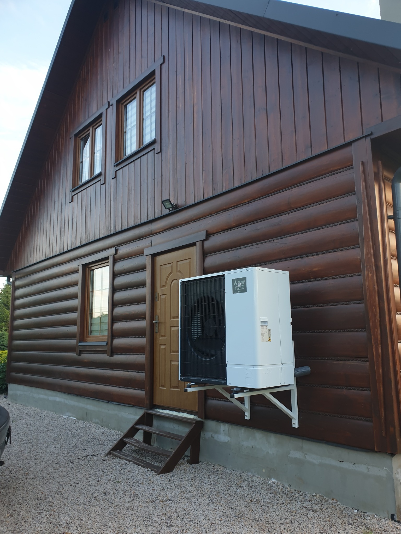 Boczny widok domu z brązowych bali z zamontowaną białą pompą ciepła Mitsubishi na wspornikach. Widoczne okna, drzwi wejściowe i kamieniste podłoże.