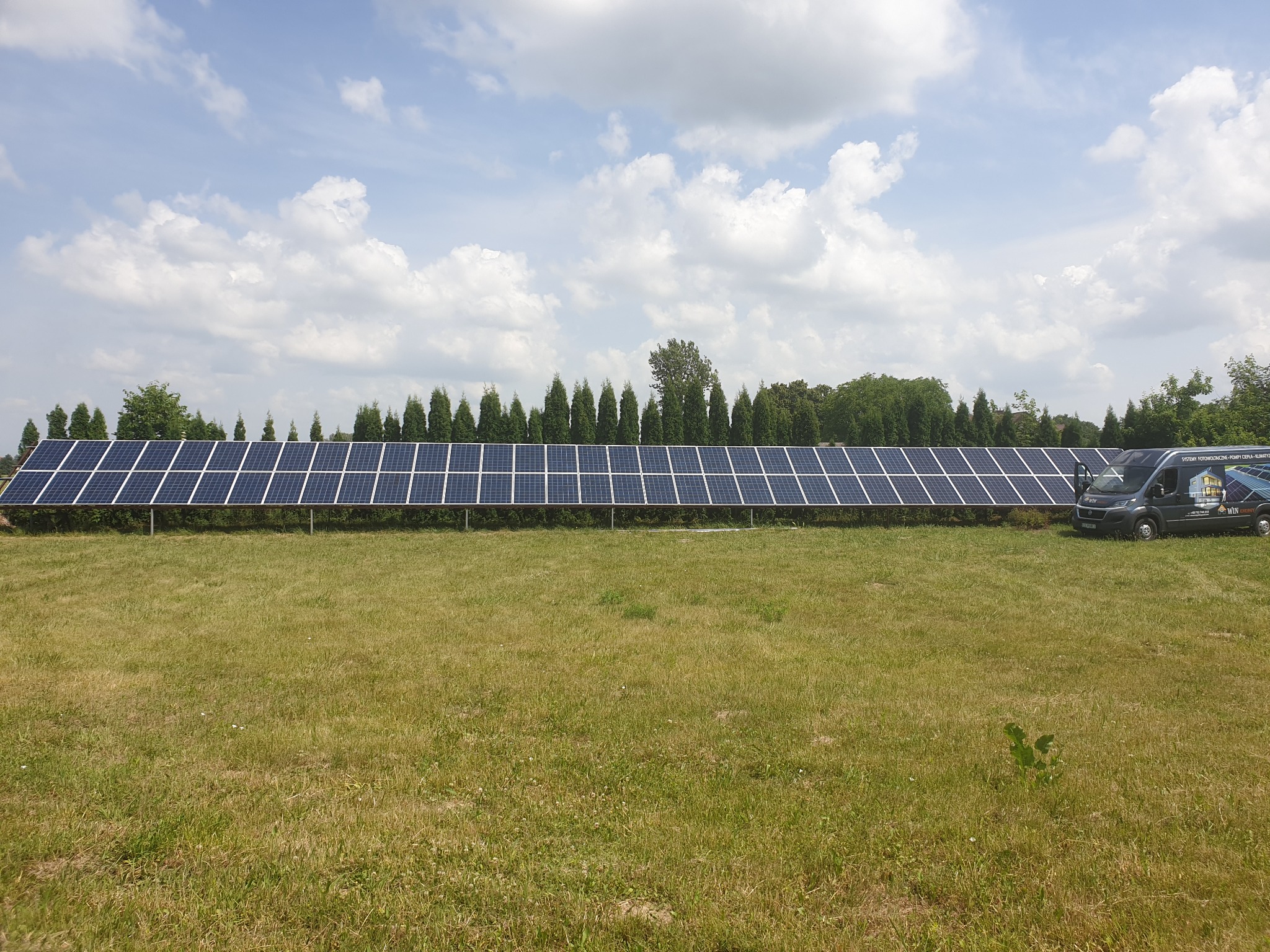Długi rząd paneli fotowoltaicznych na zielonej łące pod błękitnym niebem z chmurami. Obok zaparkowany bus z logo firmy. Widok z poziomu gruntu.