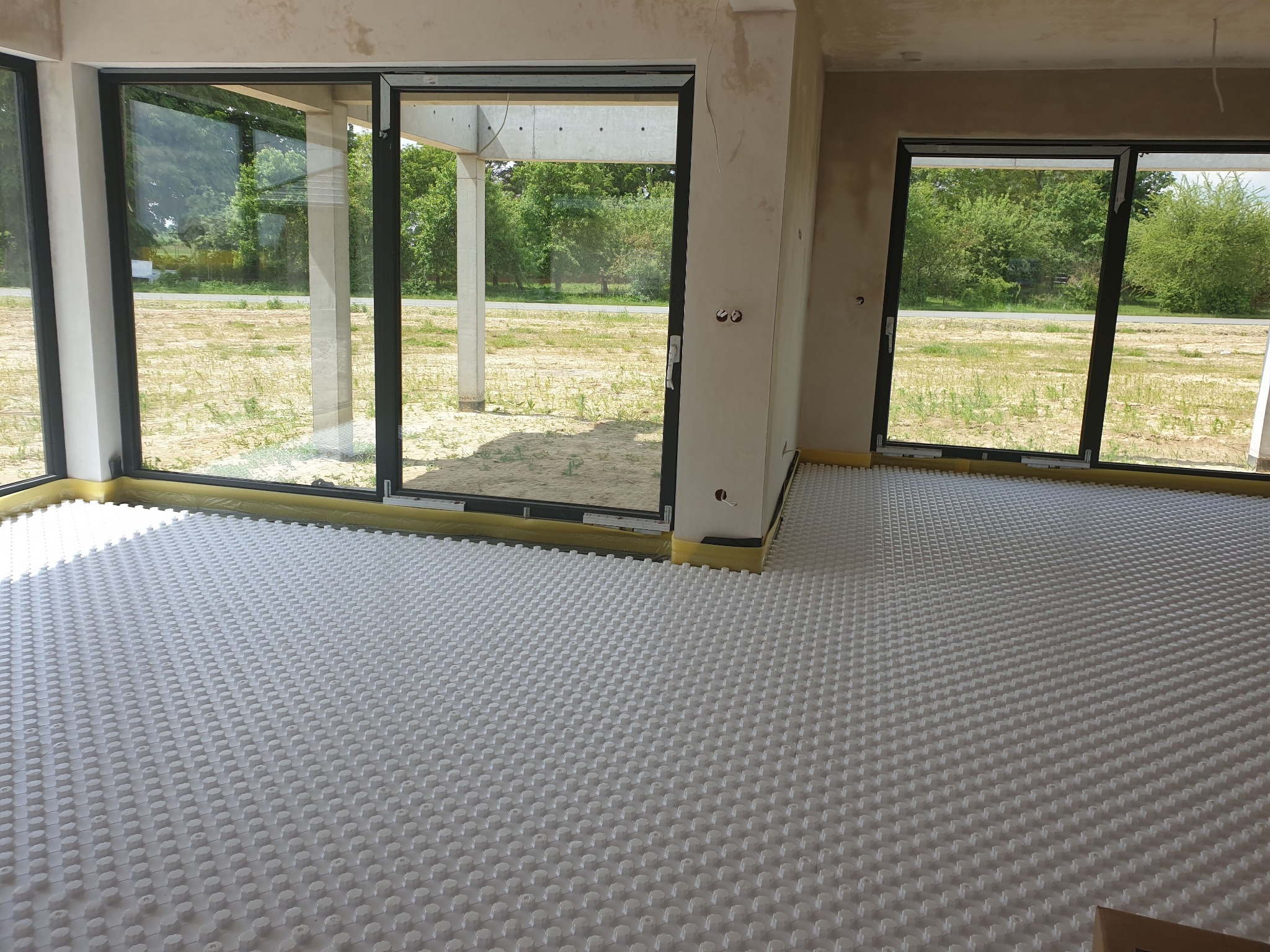 Biała mata do ogrzewania podłogowego z widocznymi otworami na rury, rozłożona w surowym wnętrzu z dużymi oknami, przez które widać zieleń.