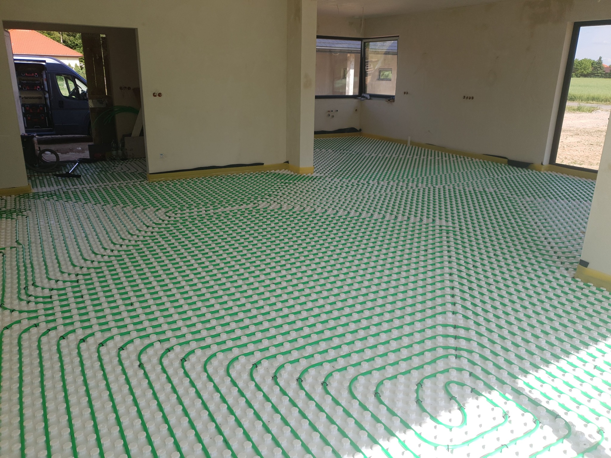 Ułożone rury ogrzewania podłogowego w jasnym pomieszczeniu, widok z góry. Na drugim planie widoczny samochód dostawczy z narzędziami.