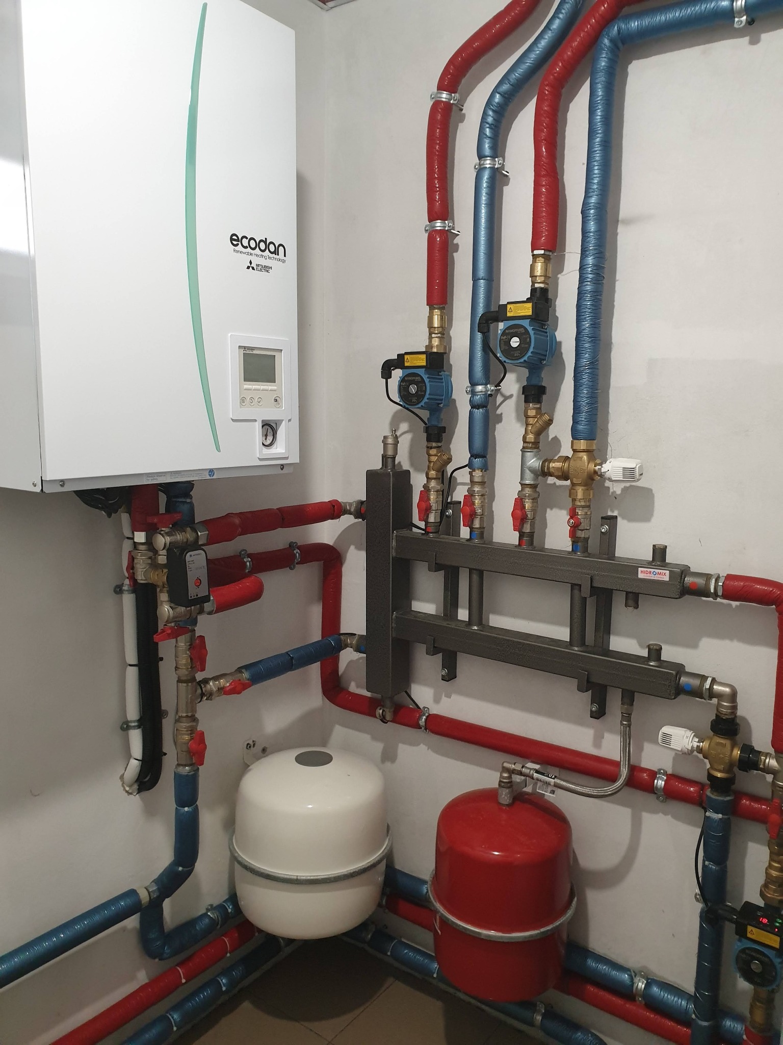 Instalacja pompy ciepła Ecodan z widocznymi rurami w izolacji czerwonej i niebieskiej, zbiornikami wyrównawczymi i pompami obiegowymi w pomieszczeniu technicznym.