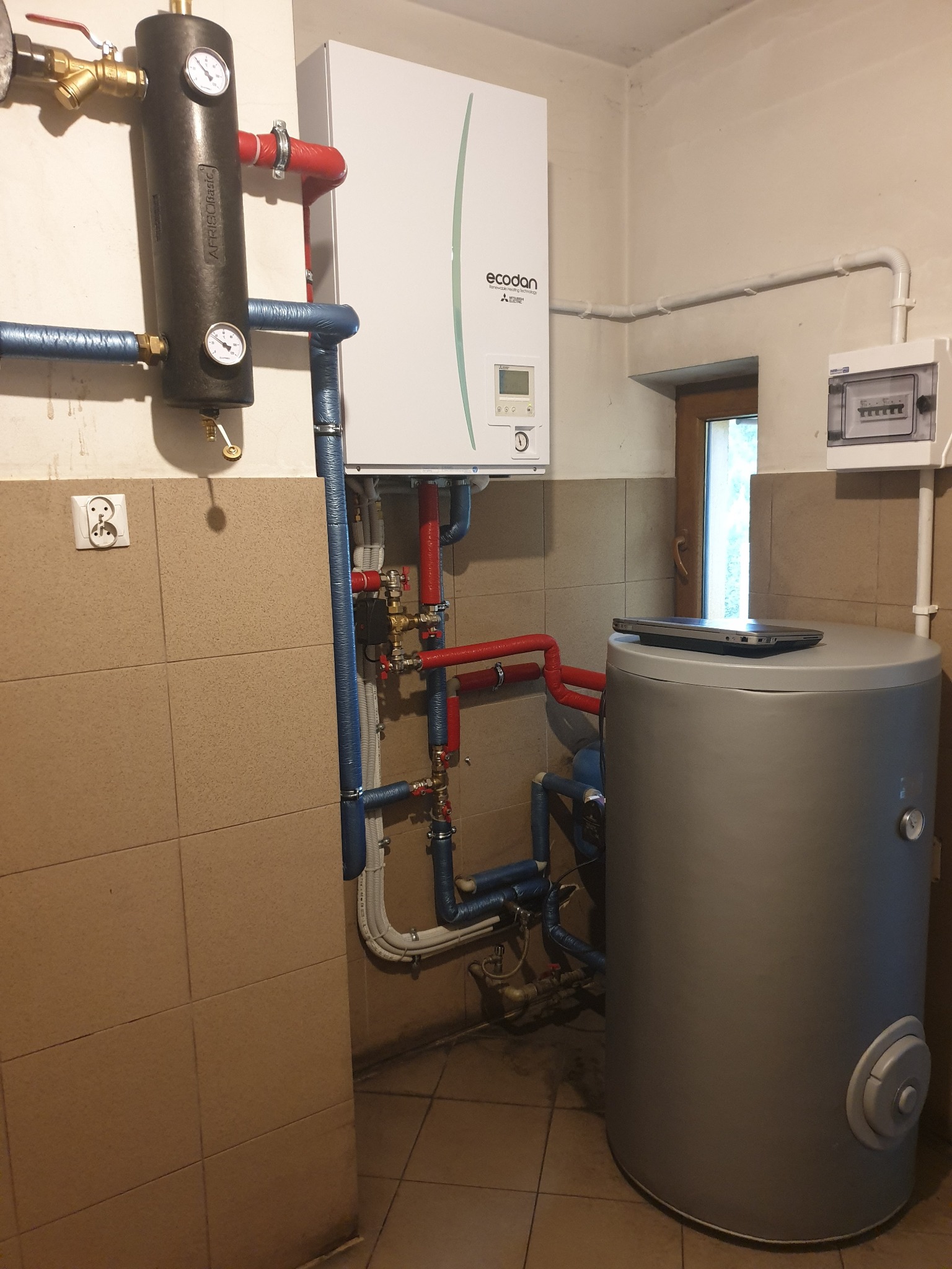 Zmontowana instalacja pompy ciepła Ecodan z zasobnikiem, rurami w izolacji i czarnym separatorem powietrza. Na zasobniku leży laptop. Tło to kafelki i okno.