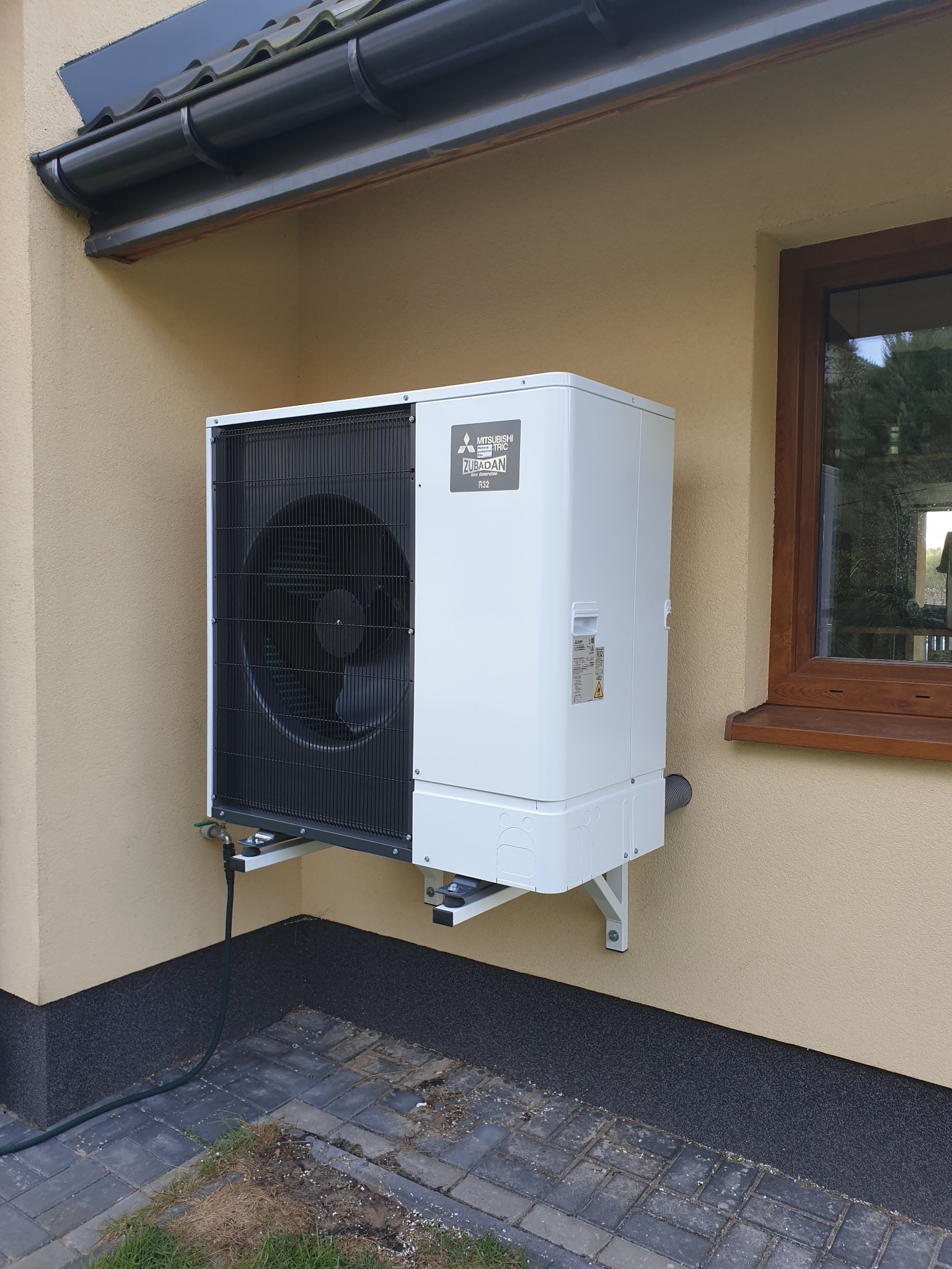 Biała, zewnętrzna jednostka pompy ciepła Mitsubishi Zubadan R32 zamontowana na elewacji budynku z beżowym tynkiem, widoczna z boku, z fragmentem okna i rynny.