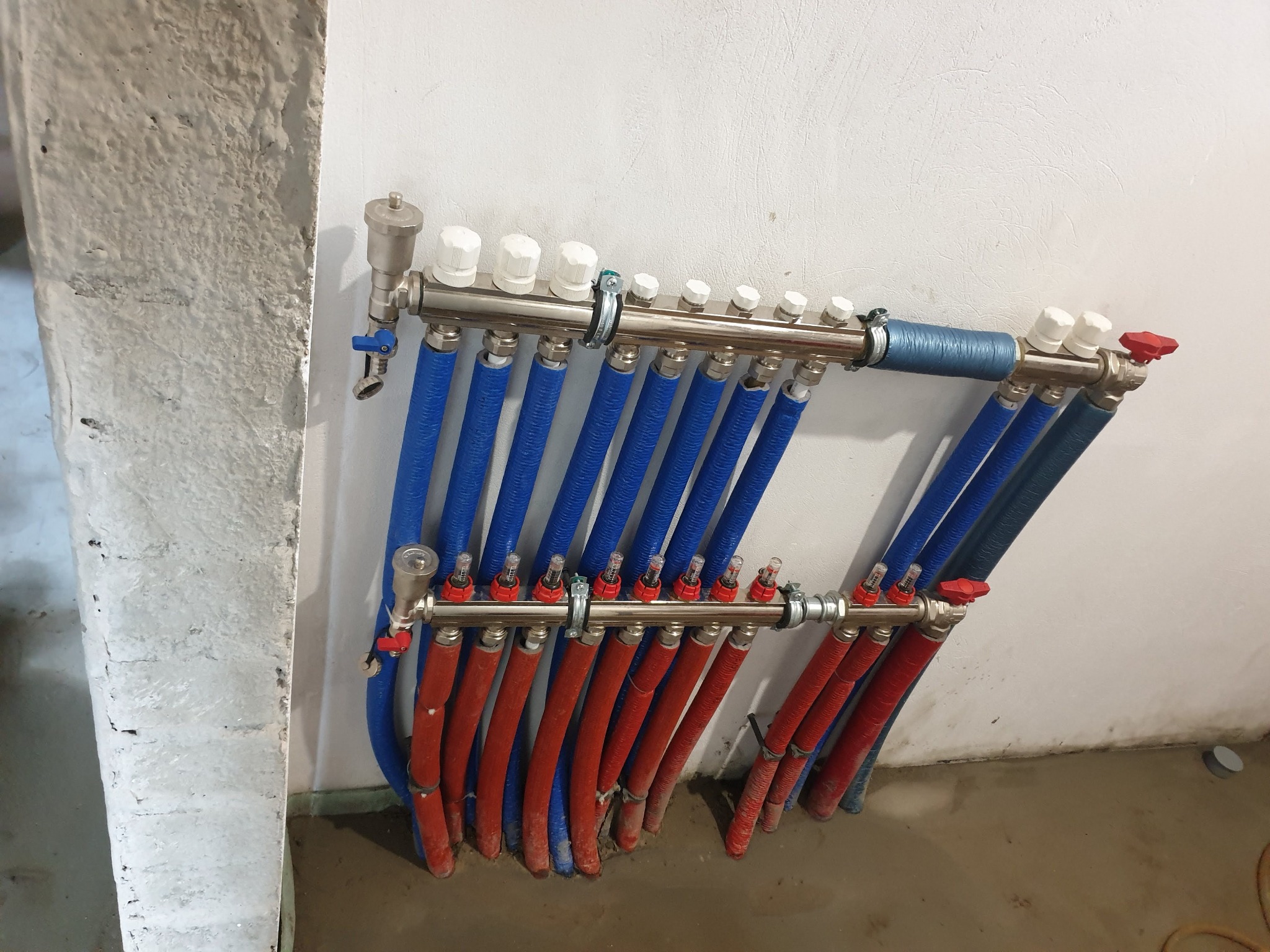 Rozdzielacz ogrzewania podłogowego z rurami w kolorach niebieskim i czerwonym, zamontowany na białej ścianie w surowym wnętrzu. Widoczne elementy regulacyjne i przyłączeniowe.