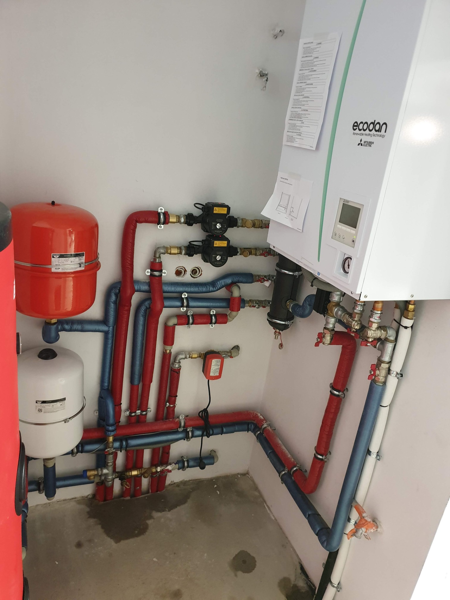 Instalacja grzewcza z piecem Ecodan, zbiornikami i rurami w izolacji czerwono-niebieskiej. Widoczne zawory i elementy montażowe. Całość w jasnym pomieszczeniu.