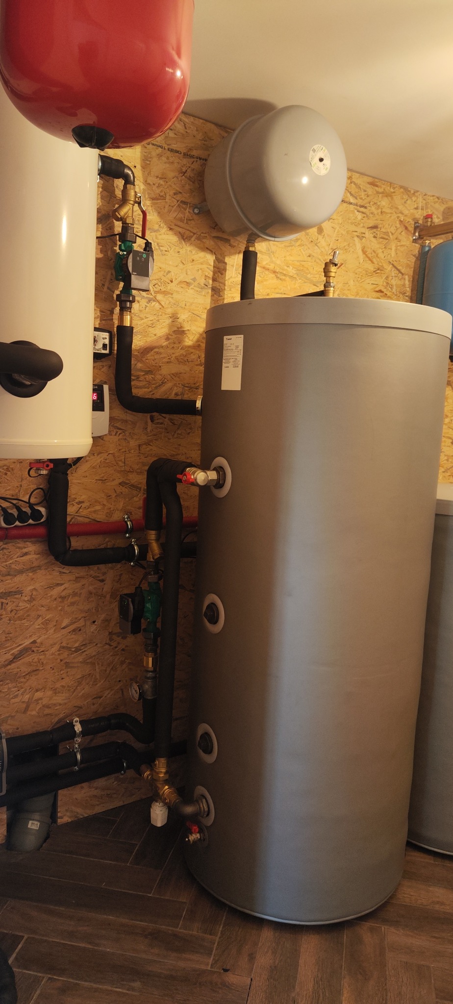 Instalacja CO z zbiornikiem buforowym, rurami w izolacji i zaworami, zamontowana na ścianie z boazerii. Widoczne elementy instalacji grzewczej.