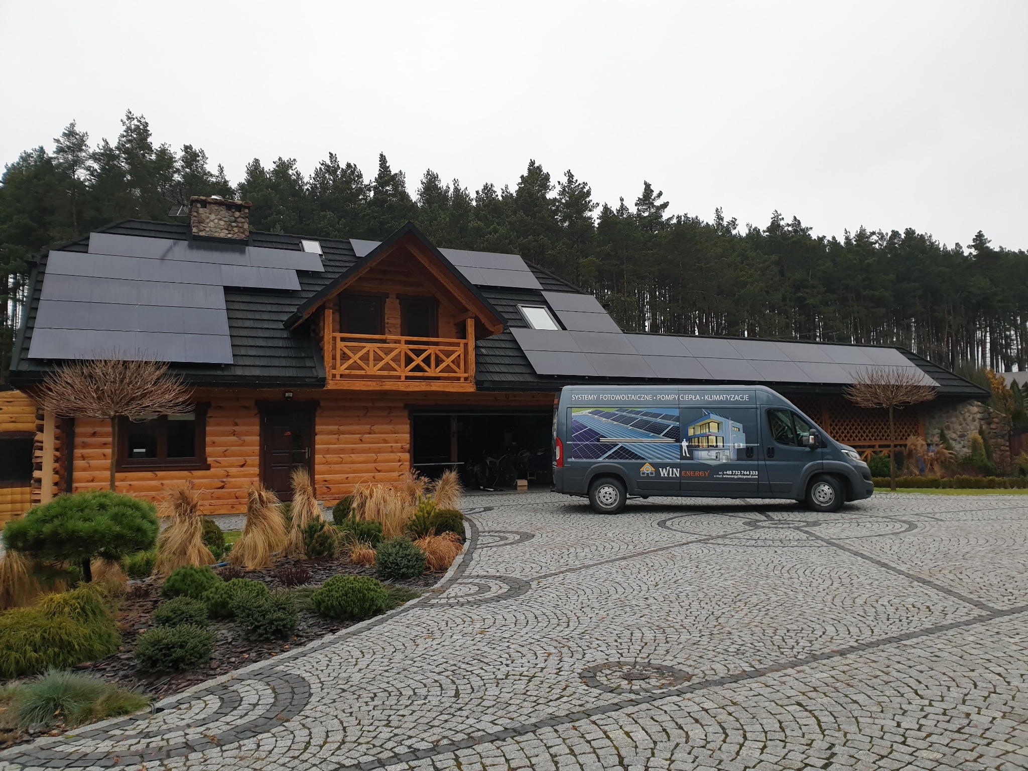 Dom z bali z panelami fotowoltaicznymi na dachu, na podjeździe stoi bus firmy instalującej systemy fotowoltaiczne, pompy ciepła i klimatyzacje.