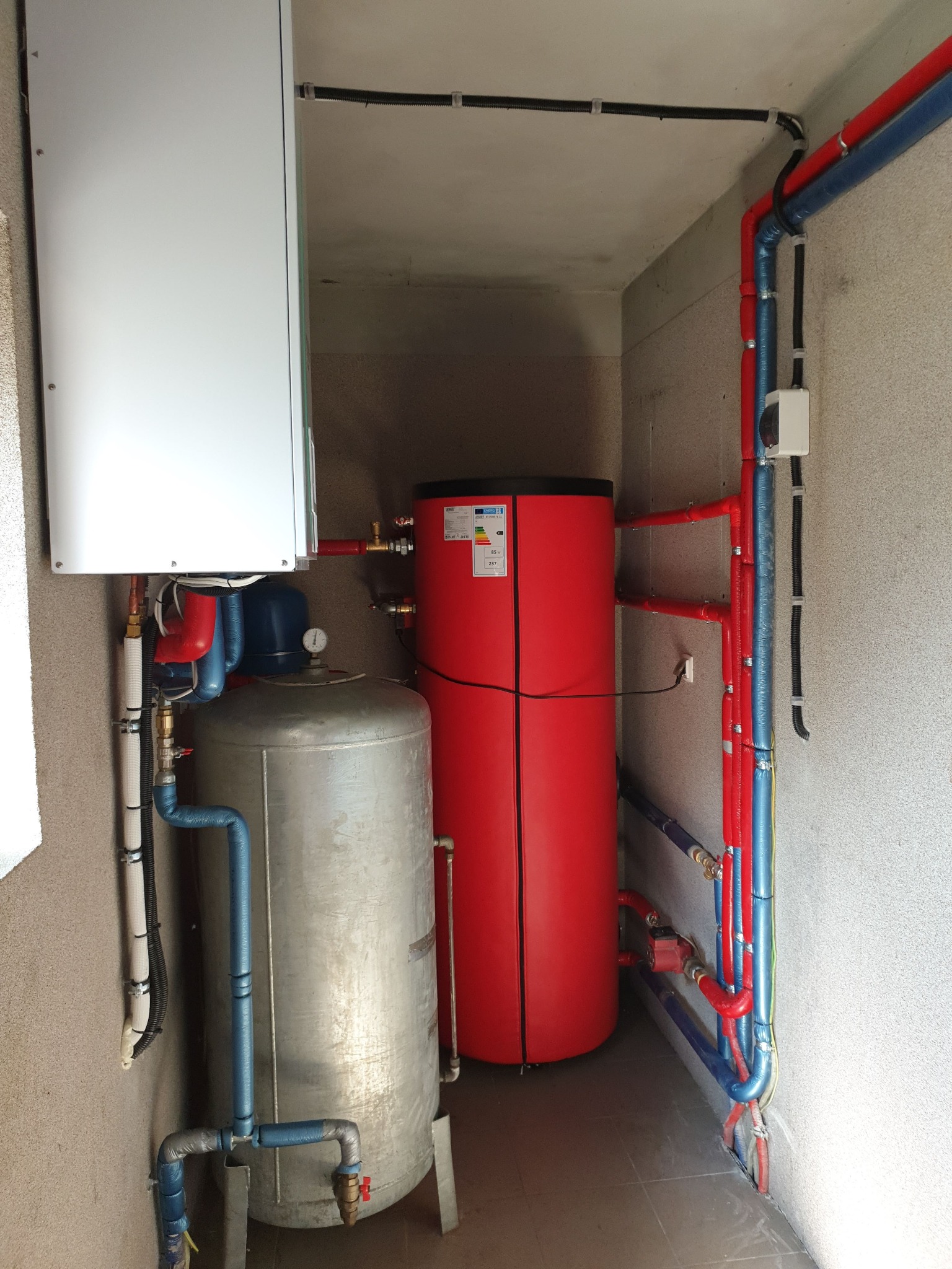 Zestaw instalacji grzewczej w kotłowni: srebrny zbiornik buforowy, czerwony zasobnik ciepłej wody i biały piec, połączone rurami w izolacji czerwonej i niebieskiej.