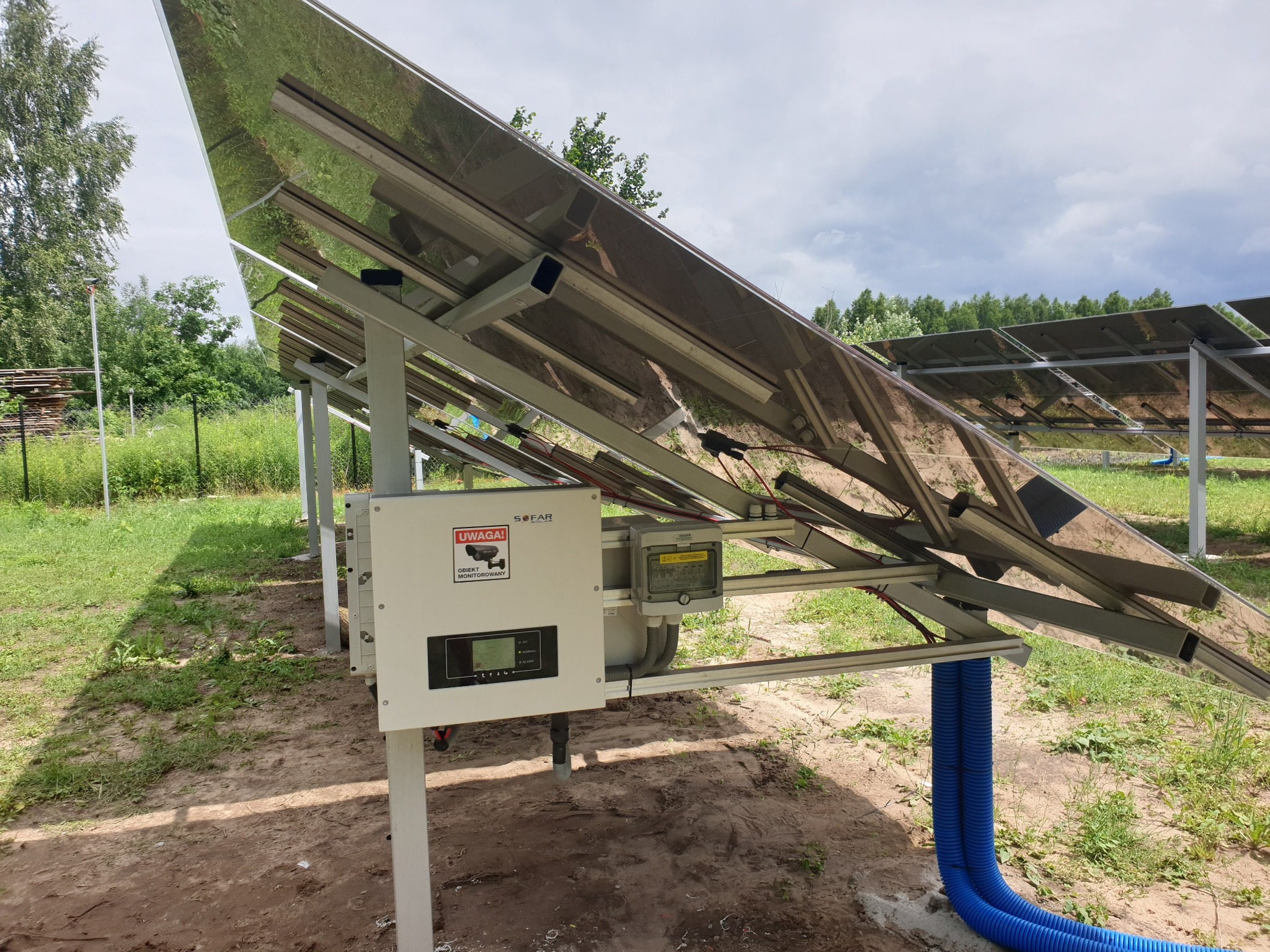 Instalacja fotowoltaiczna z widocznym inwerterem i niebieskimi rurami osłonowymi na tle zieleni. Panele odbijają otoczenie, tworząc lustrzany efekt.