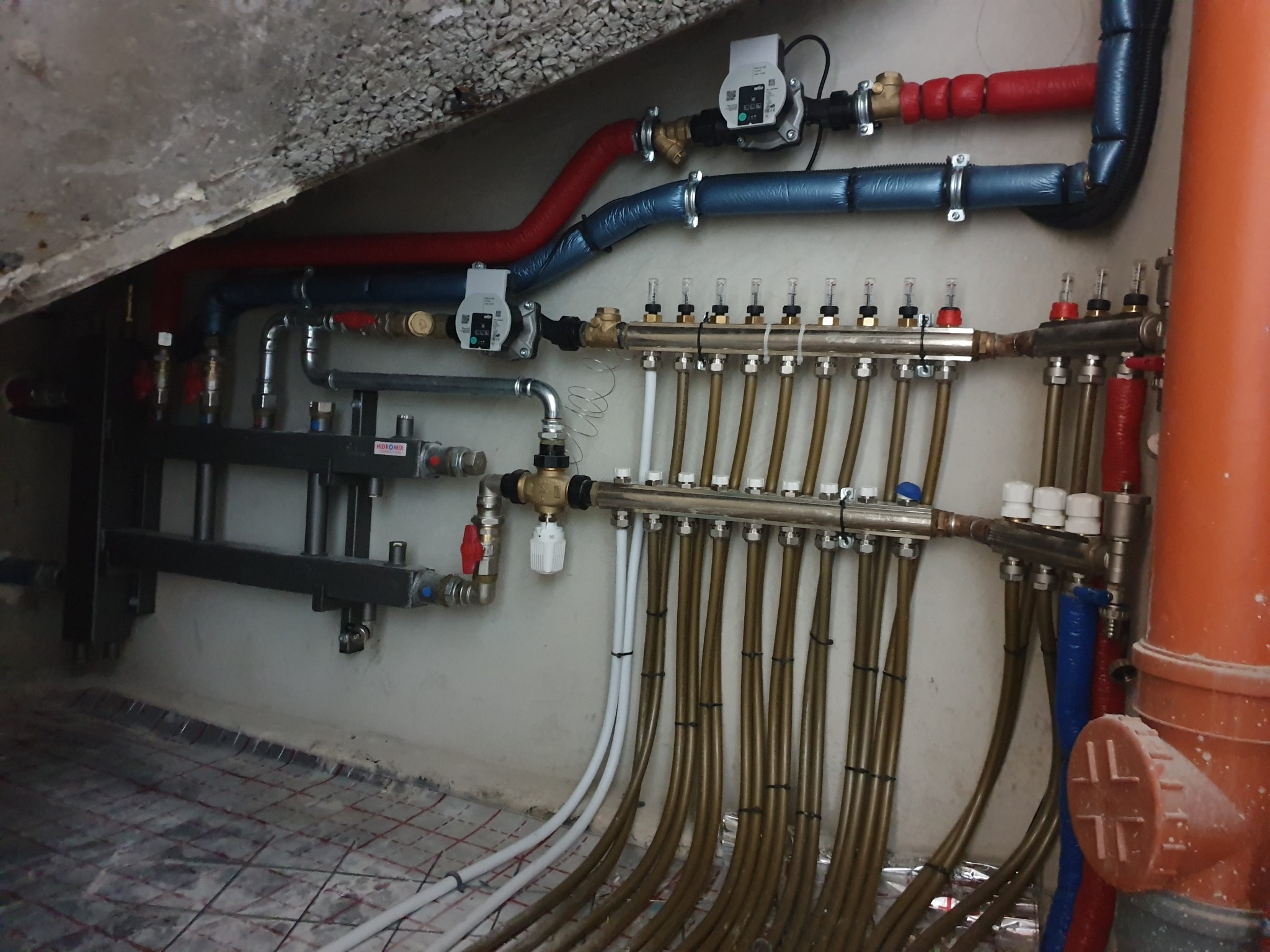 Rozdzielacz ogrzewania podłogowego z licznymi rurami, zaworami i elementami sterującymi, zamontowany na ścianie w pomieszczeniu z widoczną siatką grzewczą w podłodze.
