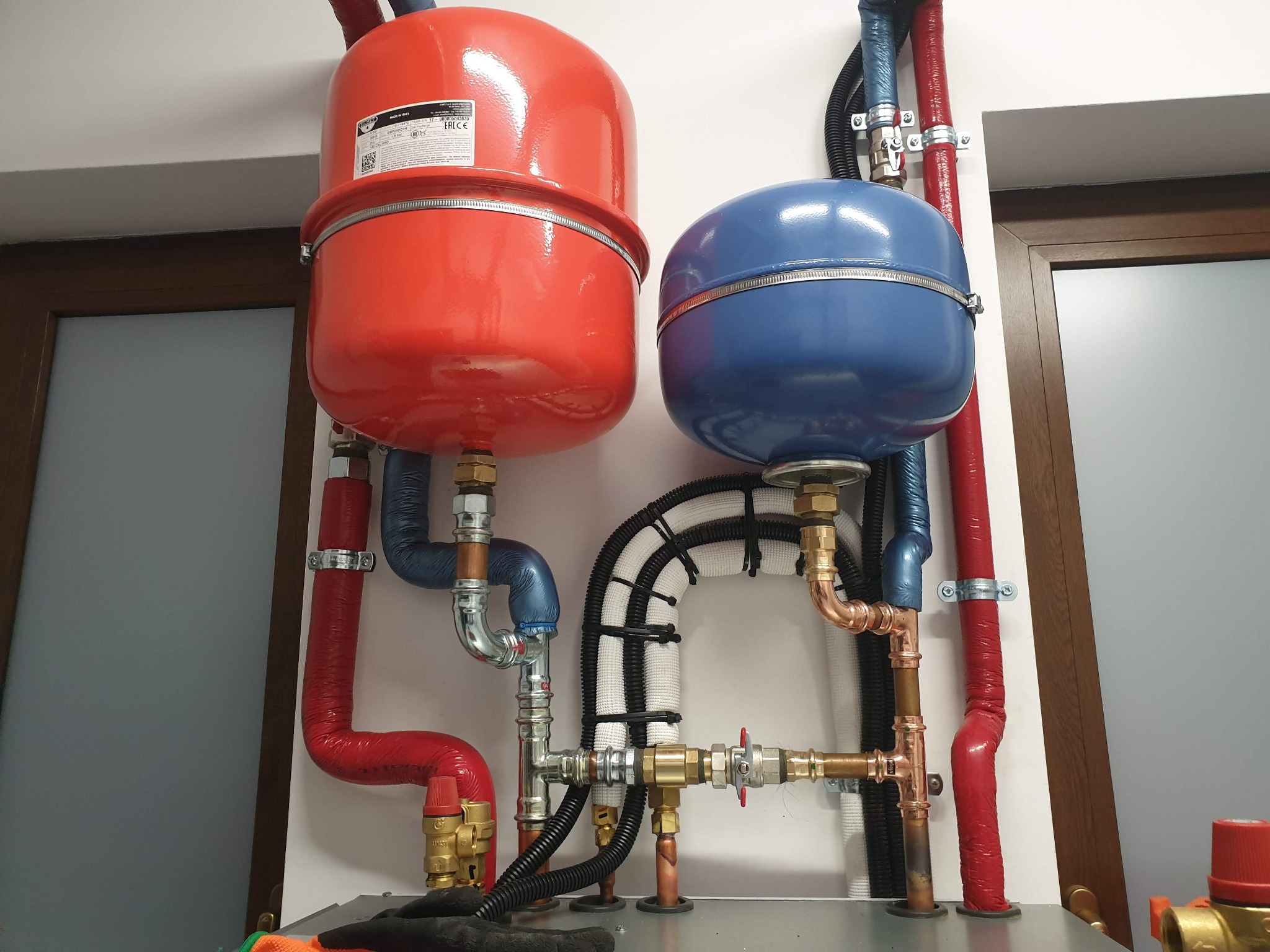 Instalacja CO z czerwonym i niebieskim zbiornikiem wyrównawczym, rurami w izolacji i zaworami, na tle białej ściany i fragmentu okna.
