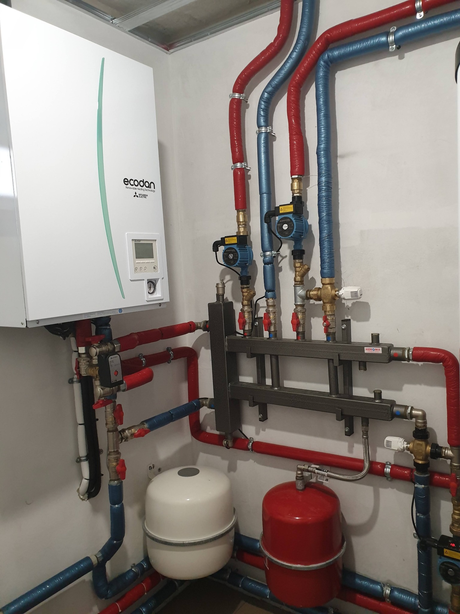 Biała pompa ciepła Ecodan i instalacja hydrauliczna z rurami w kolorze czerwonym i niebieskim w Ciecierzynie. Widoczne zawory i zbiorniki.
