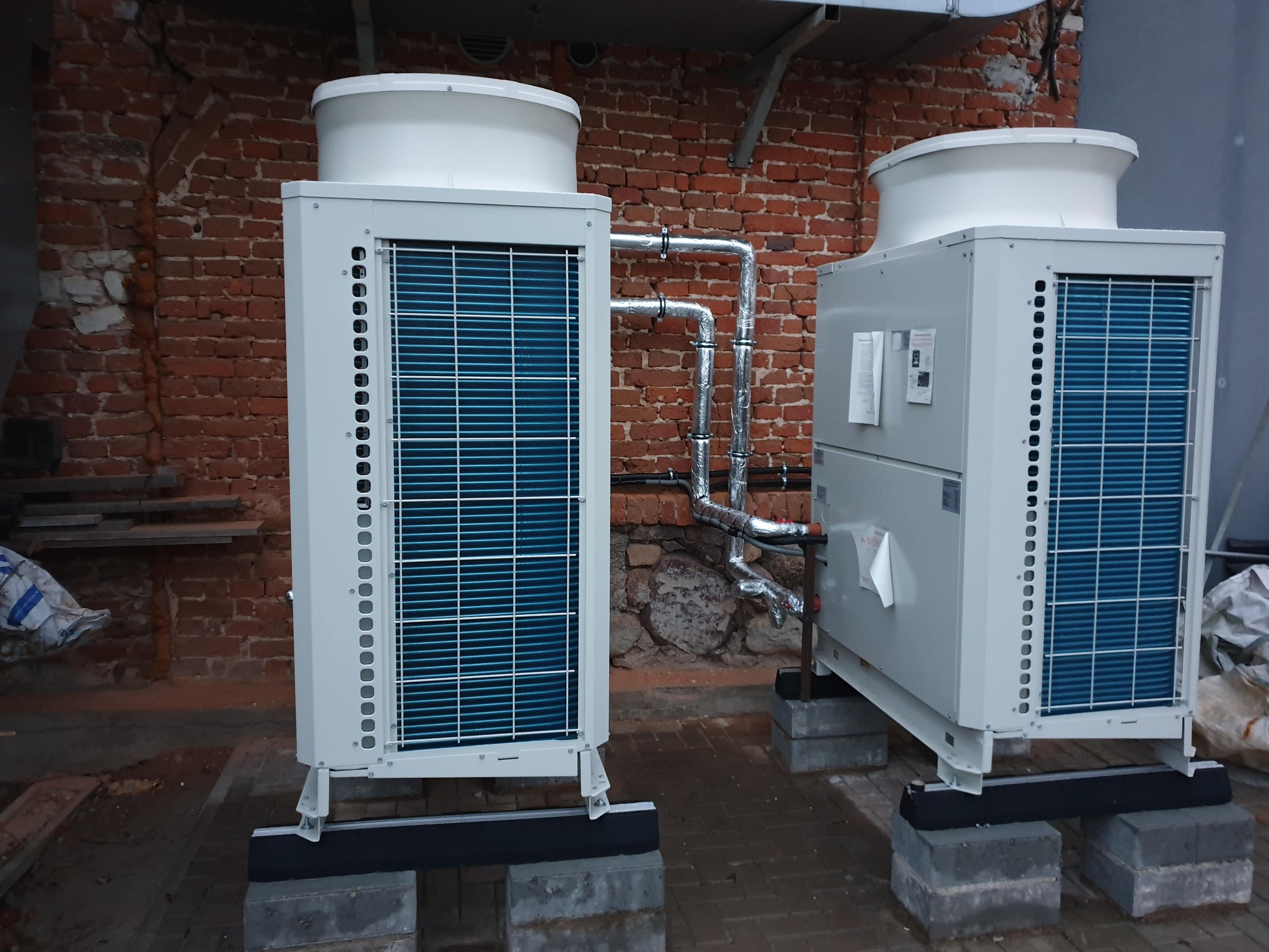 Dwie białe pompy ciepła z niebieskimi lamelami, połączone rurami, stoją na betonowych blokach na tle ceglanej ściany. Widoczne elementy instalacji i otoczenia.