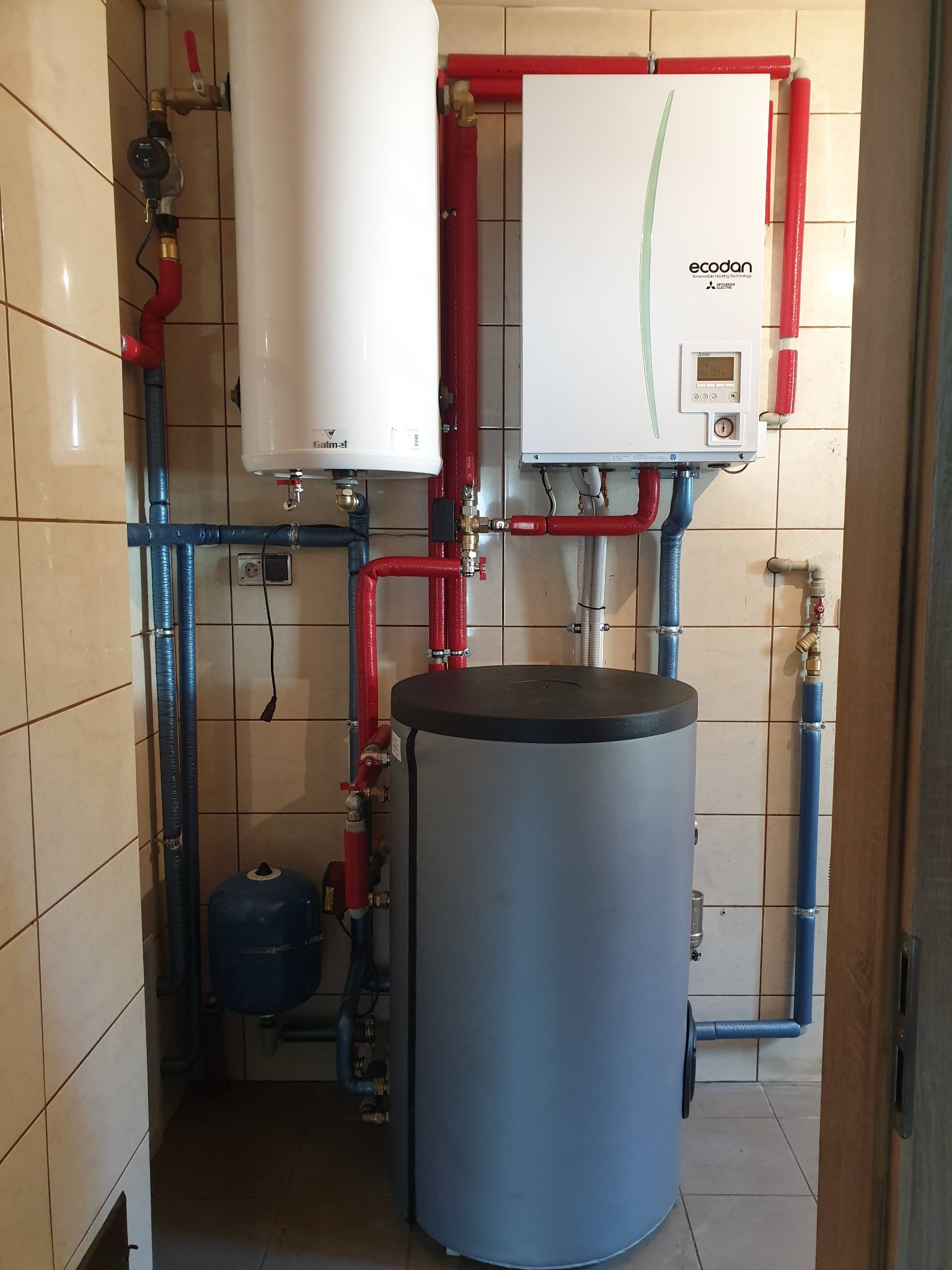 Zestaw instalacji grzewczej w kotłowni: biały zasobnik Galmet, pompa ciepła Ecodan Mitsubishi i szary zbiornik akumulacyjny, połączone rurami w izolacji.
