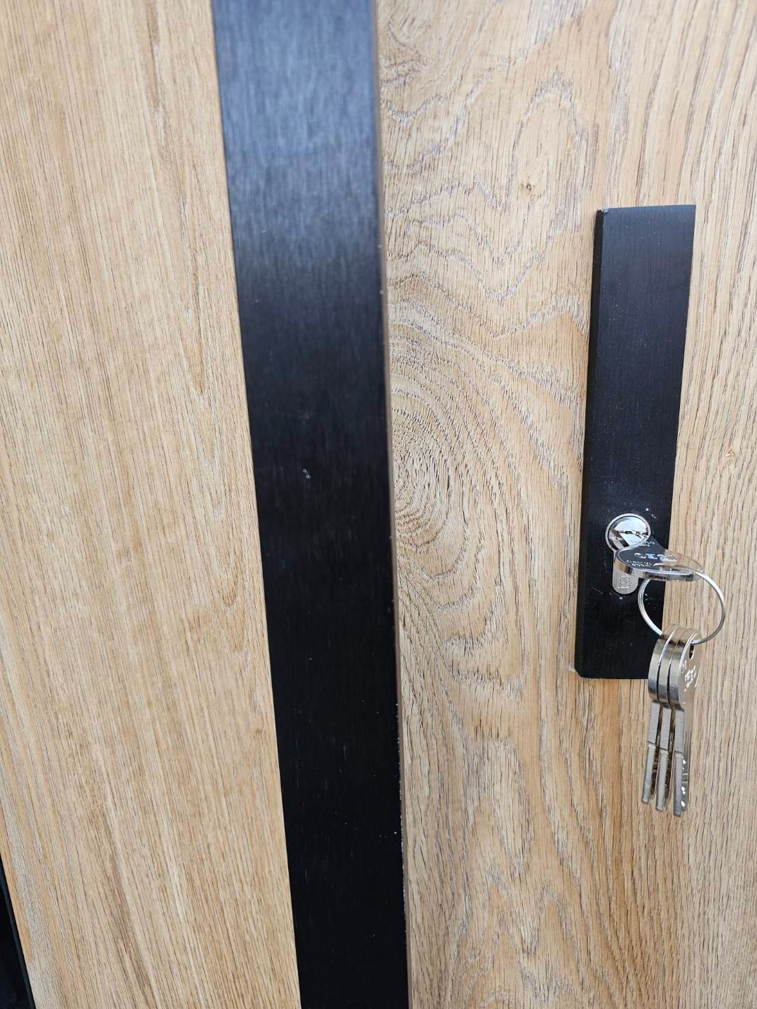 Klucze w zamku nowoczesnych drzwi z drewnianą okleiną i czarnymi elementami dekoracyjnymi. Detal zamka i kluczy.