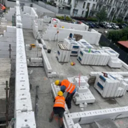 Wznoszenie ścian z białych bloczków silikatowych na budowie. Dwóch pracowników w kaskach i kamizelkach układa elementy. Widok z góry na powstającą konstrukcję.