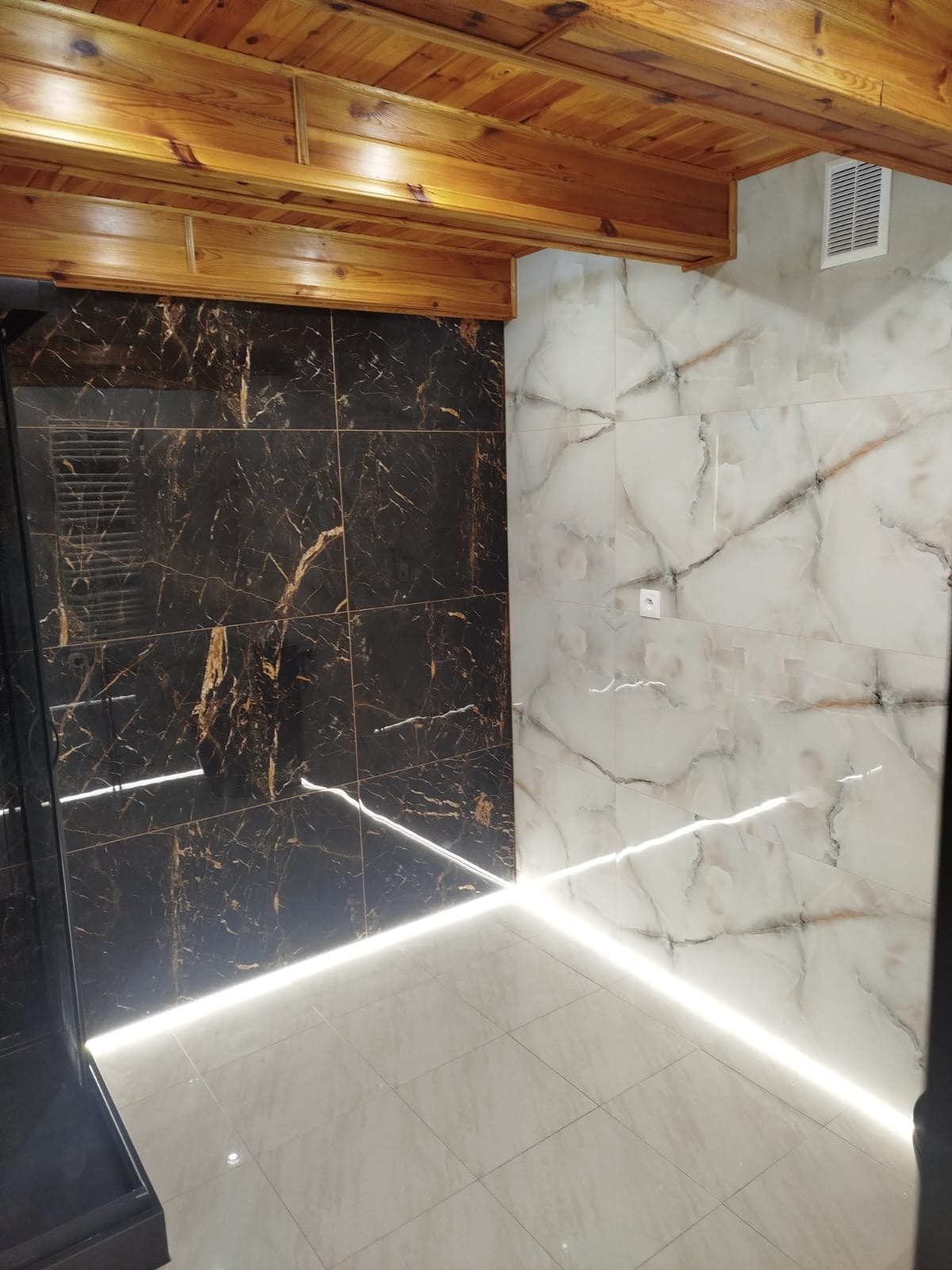 Wykończona łazienka z czarno-złotymi i białymi marmurowymi płytkami, podświetleniem LED przy podłodze i drewnianym sufitem. Nowoczesny design z elementami rustykalnymi.