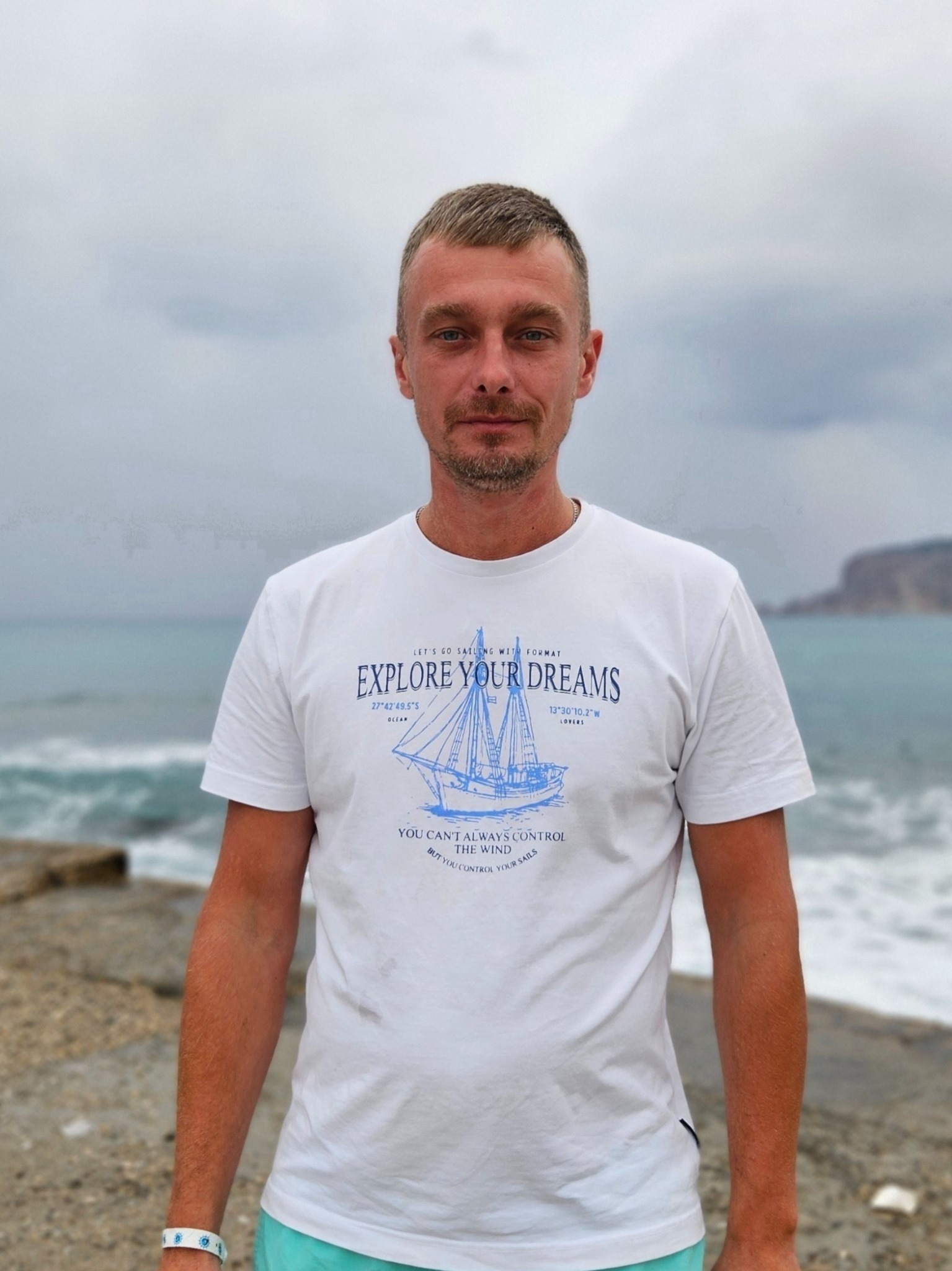 Mężczyzna w białej koszulce z nadrukiem żaglówki na tle morza i pochmurnego nieba. Ujęcie portretowe, z widoczną plażą i klifem w oddali.