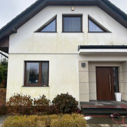 Maciej Ellwart - Biała elewacja domu z widocznymi zabrudzeniami i oknami o brązowych ramach. Na pierwszym planie widoczny wąż ciśnieniowy sugerujący przygotowania do mycia.