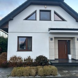 Maciej Ellwart - Biały dom jednorodzinny z ciemnym dachem i brązowymi oknami, widok z poziomu chodnika. Widoczne krzewy przed budynkiem i fragment chodnika z kostki.