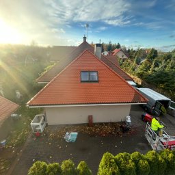 Maciej Ellwart - Czerwony dach domu jednorodzinnego z okienkiem dachowym, widok z góry. Pracownik na podnośniku koszowym, sprzęt do czyszczenia, jesienne liście na kostce brukowej.