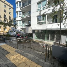 Maciej Ellwart - Pracownik na podnośniku koszowym czyści elewację nowoczesnego budynku z balkonami. Widoczne okna i szyldy firm. Ujęcie z poziomu chodnika z kostki brukowej.