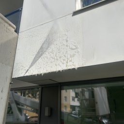 Maciej Ellwart - Mycie elewacji budynku z podnośnika koszowego. Strumień wody usuwa zabrudzenia, tworząc efekt czyszczenia na jasnej fasadzie. Widoczne okna i odbicie otoczenia.