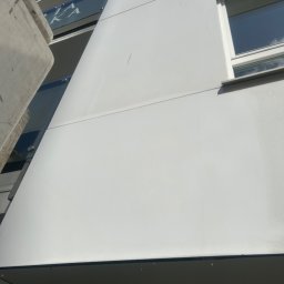 Maciej Ellwart - Biała elewacja budynku po umyciu, widoczne krople wody, okno z białymi ramami, fragment balkonu z szklaną balustradą. Ujęcie z dołu, podkreślające wysokość budynku.