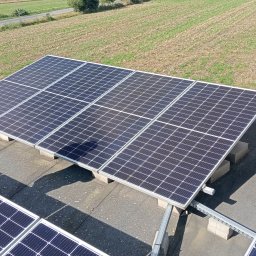 Piotr Napierała - Panel słoneczny na dachu z widoczną osobą. Ujęcie z góry, ukazujące instalację fotowoltaiczną na tle zielonego pola. Widoczne elementy montażowe paneli.