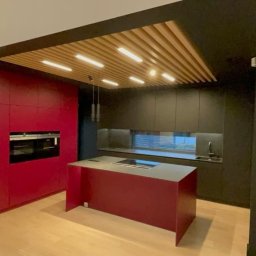 SPOTLESS RAFAŁ MICHALUK - Nowoczesna kuchnia z wyspą, czerwonymi szafkami i drewnianym sufitem z oświetleniem. Minimalistyczny design, jasna podłoga, czarne ściany i szafki.