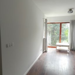 SPOTLESS RAFAŁ MICHALUK - Jasne, świeżo pomalowane wnętrze pokoju z drewnianą podłogą i wyjściem na balkon. Widoczne gniazdka elektryczne, włączniki światła i minimalistyczna lampa sufitowa.