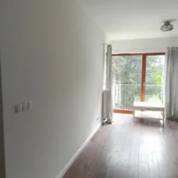 Jasne, świeżo pomalowane wnętrze pokoju z drewnianą podłogą i wyjściem na balkon. Widoczne gniazdka elektryczne, włączniki światła i minimalistyczna lampa sufitowa.