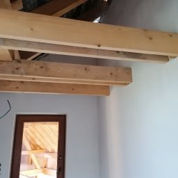Tynki Spider - Poddasze w trakcie ocieplania: drewniana konstrukcja stropu, widoczna izolacja, fragment ściany i okno. Prace wykończeniowe w jasnym wnętrzu.