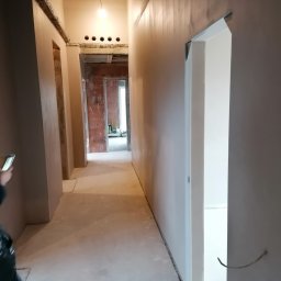 Tynki Spider - Korytarz w trakcie prac wykończeniowych: ściany pokryte gładzią, widoczne otwory na oświetlenie, surowy beton na podłodze, częściowo zamontowane ościeżnice.